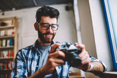 Başarılı hippi erkek bekleyen arkadaş fotoğraflar vintage fotoğraf makinesi, serbest zaman sırasında kontrol ederken kitabevi içinde mutlu şık gülümseyen adam eski moda ekipman tutan trendy giyim giymek