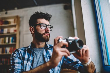 Düşünceli hipster adam giyim gözlük vizyon düzeltme imge yolu ile retro kamera, konsantre profesyonel erkek fotoğrafçı zevk zaman ve hobi kapalı yaptıktan sonra Vitrinlere bakıyor