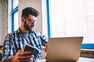Cep telefonu, modern dizüstü bilgisayar ve bekleyen arkadaş kapalı ile vakit Üniversitesi Kampüsü oturan ciddi hipster adam tutarak şaşkın erkek profesyonel metin yazarı konsantre
