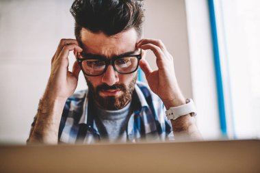 Genç beyaz erkek programcı son tarih içinde yazıhane bilgisayar, şaşkın şaşkın hipster adam kötü haber için wifi bağlıyken modern netbook üzerinde okuma ile deckstop oturan baş ağrısı hissediyor