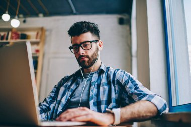 Bilgi için modern teknolojiyi kullanarak ve sesli kitap ciddi hipster adam gözlük netbook üzerinde Internet tarama yolu ile kulaklıklar, dinleme yeni makale arama erkek metin yazarı konsantre