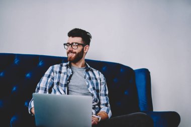 Modern dizüstü bilgisayar ile rahat kanepe oturma ve e öğrenme, başarılı hipster adam Eyewear bir kenara netbook üzerinde çalışmanın tatilinde yüzünde sevimli gülümseme ile seyir zevk mutlu beyaz blogger