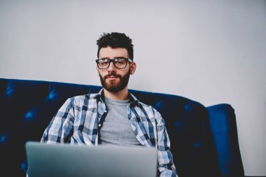 Netbook ile rahat kanepe oturma ve kamera, dizüstü bilgisayar üzerinde çalışan tatilinde poz trendy Gözlük Genç beyaz hipster adam bakarak yakışıklı bin yıllık hipster adam portresi 