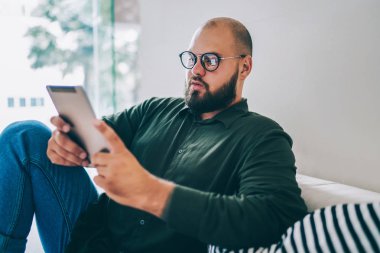 Dalgın adam gözlük vizyon düzeltme için dijital tablet, ciddi erkek blogger kontrol mesajları ve boş olarak Wi-Fi bağlantısı kullanarak ağ güncelleştirmeleri yeni app yükleme üzerinde konsantre