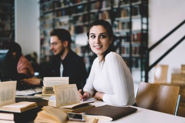 Ödev eğitim edebiyat ve ders kitapları, ders projesi Üniversite Kütüphane içinde okul sırası, oturmak için araştırma yapma olumlu genç kadın kullanarak üniversite kampüsü içinde gülümseyen hippi kız