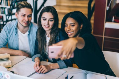 Neşeli çok ırklı mürettebat smartphone fotoğraf makinesinde yaygın fotoğraf için poz serbest zaman sırasında öğrencilerin birlikte kampüs içinde oturan cep telefonunda selfie alan genç erkek ve dişi öğrenciler gülümseyen