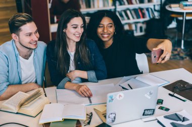 Birlikte, poz selfie hücresel togethe tarih için çok ırklı erkek ve dişi hipsters okurken arkadaşları ile resim alma ve paylaşım için akıllı telefon kullanan Afro-Amerikan Kız gülümseyerek