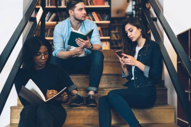 Grup çok ırklı erkek ve dişi hipsters görmezden kitap okuma ve sohbet için genç öğrenciler college Kampüsü merdivenlerde oturan boş zamanlarında harcama cep telefonuyla gerçek konuşma 