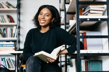 İlginç bir kitap ile memnun gülümseyen neşeli Afro-Amerikan Kız portresi bulmak Üniversite Kitaplığı'nda, Genç koyu tenli kadının boş zamanlarında kamera bakmak edebiyat okuma Kitabevi