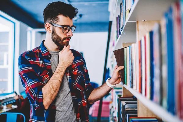 高校图书馆眼镜选材书中的体贴男性站在书架附近 集中的时髦的家伙休闲打扮采摘文学和在喜爱的书店做研究 — 图库照片