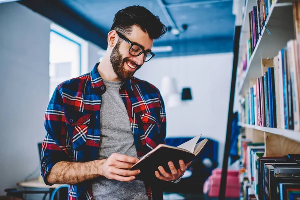 聪明的微笑的白种男性在休闲穿戴读新的有趣的小说在书店 成功的快乐的人学生拿着正面书 当站立在大学图书馆的架子附近时 — 图库照片