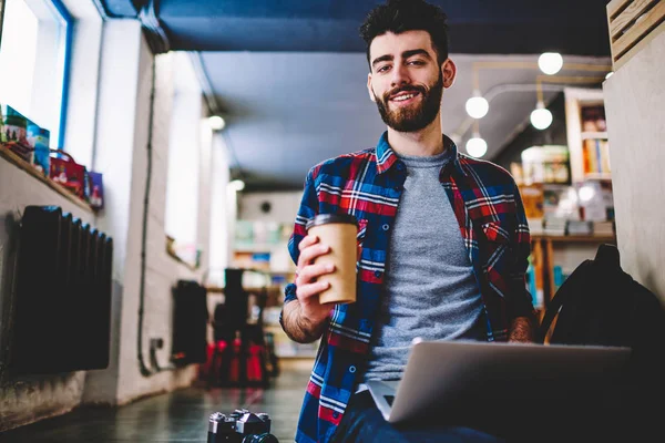Portrait of successful hipster guy enjoying morning takeaway coffee at university campus and spending time for e learning, happy male copywriter looking at camera in break of work on laptop computer
