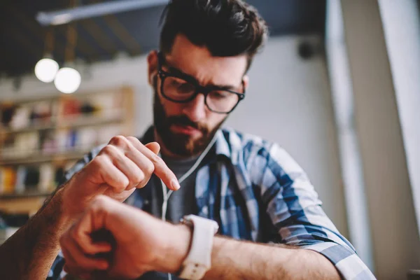 Selective focus on male hands with modern wearable smartwatch, young caucasian hipster guy checking time and gps tracking on wristwatch while enjoying new playlist via electronic earphones indoors