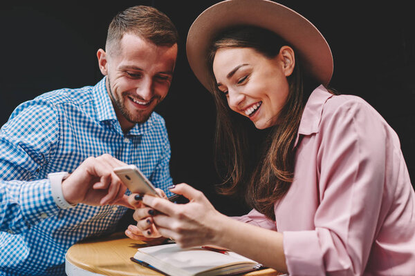Positive young marriage laughing while doing shopping online on smartphone device using free 4G internet.Cheerful stylish couple in love discussing funny video on telephone sitting in cafe interior