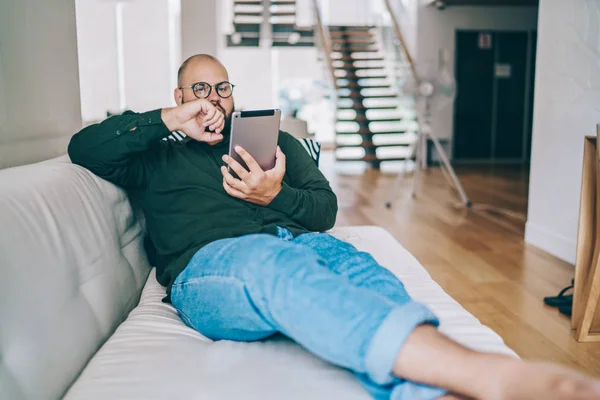 Pensive caucasian male in spectacles reading online e book via application on portable pc, hipster guy lying on sofa recreating at apartment watching movie via digital tablet and wifi at home