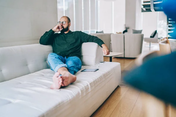 Pensive casually dressed man dreaming while sitting on white couch in modern designed living room, contemplative male blogger thinking about idea looking away resting on cozy sofa during leisure