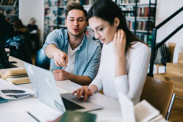 Young male student pointing on laptop computer while his female colleague typing and making research for project,clever colleagues cooperating on course work in modern library with wifi connection