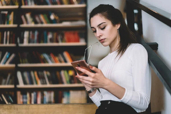 专注的年轻女性阅读智能手机的信息花在图书馆时间 沉思的女学生连接到免费无线上网在大学通过手机网络和检查邮件 — 图库照片