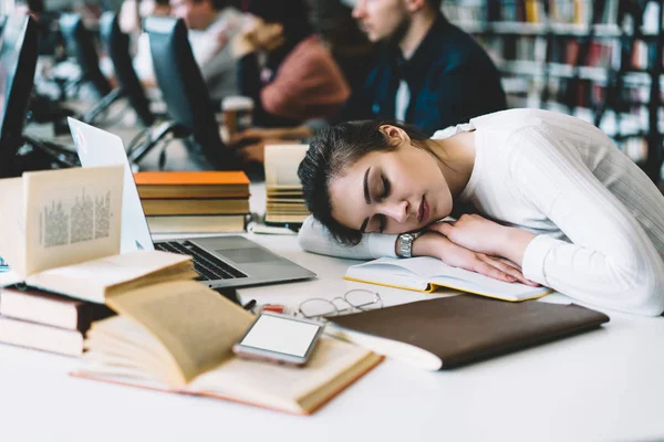 Young woman sleeping at desktop near books and literature in university campus overworked during making coursework project, smartphone with blank screen on working place, exhausted female student