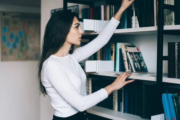 Serious young woman in casual wear standing near bookshelves picking literature for education in college,female librarian ordering books for picking concentrated on job, thoughtful girl choosing novel