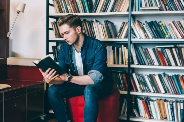 Ernster Mann Freizeitkleidung Der Neben Einem Bücherregal Sitzt Und Sich — Stockfoto