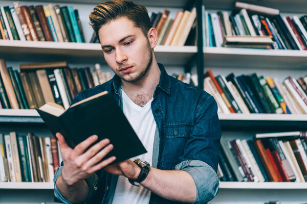 Pensive caucasian male reading interesting best seller in bookstore, serious hipster guy using textbook for getting information and learning, concentrated clever man holding literature for stud