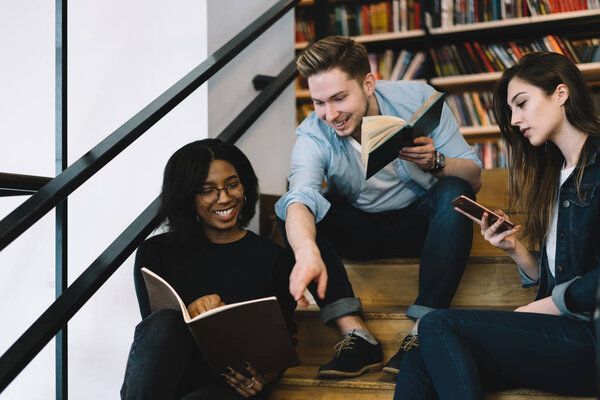 Cheerful male and female students joking and having fun while reading books in college library, woman checking mail on smartphone ignore conversation with positive friends laughing on break