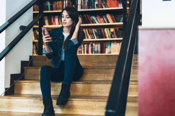 Young female in casual wear spending free time in library using wifi for blogging and chatting with friends online, hipster girl checking new feeds in social networks via app on mobile phone indoors