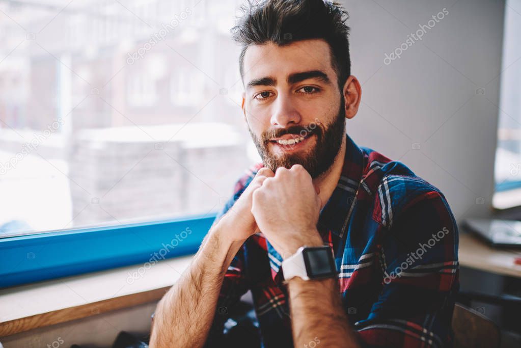 Handsome bearded positive hipster guy looking at camera with cute smile on face while waiting friend at university cafeteria, trendy successful male student wear in casual look sitting at campus