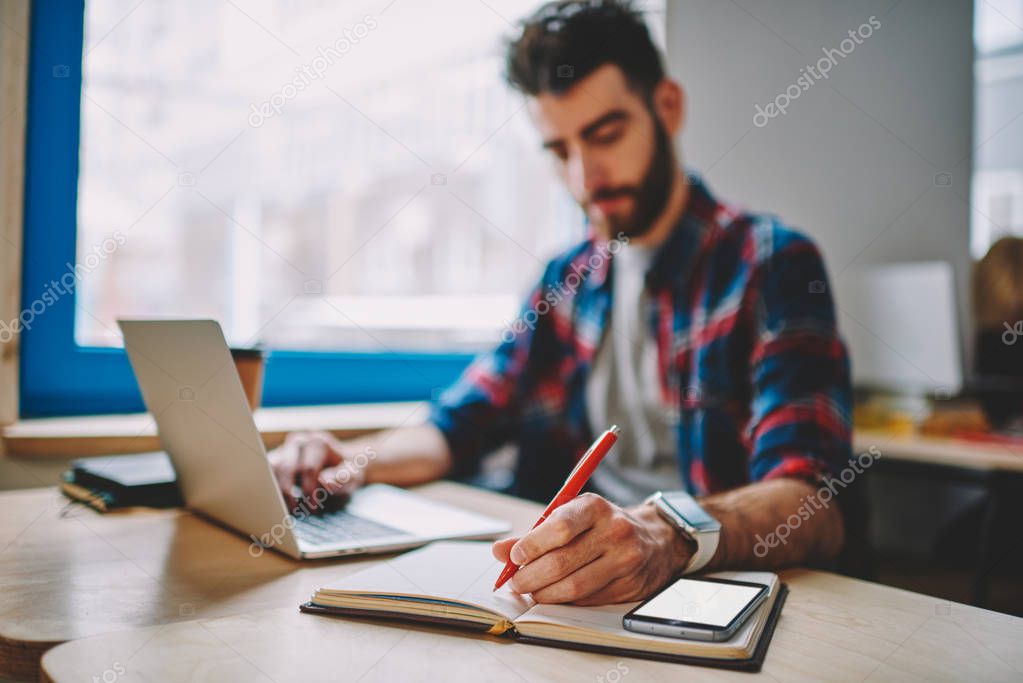 Selective focus on textbook and hand with pen,  clever male student studying indoors using modern technology and preparing for exam, smart man it developer writing organization plan in notebook