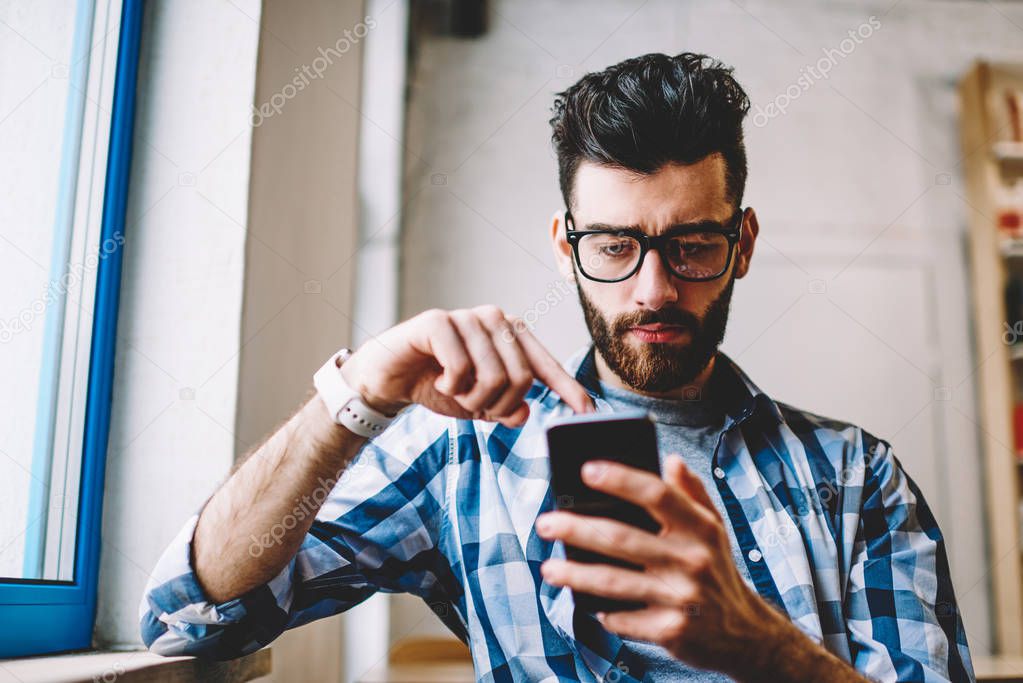 Young concentrated man making money transaction via application on mobile phone with 4g internet sitting at university campus, serious millennial man in eyewear reading text message on smartphone