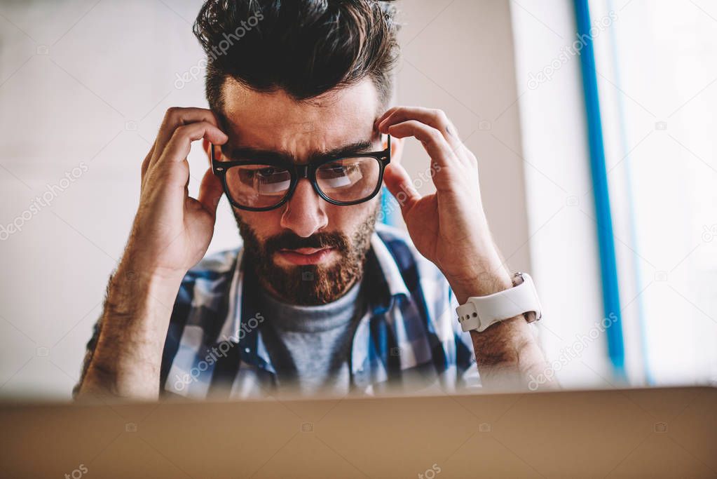 Young caucasian male programmer feeling headache from deadline sitting deckstop in office with computer, puzzled confused hipster guy reading bad news on modern netbook while connected to wifi