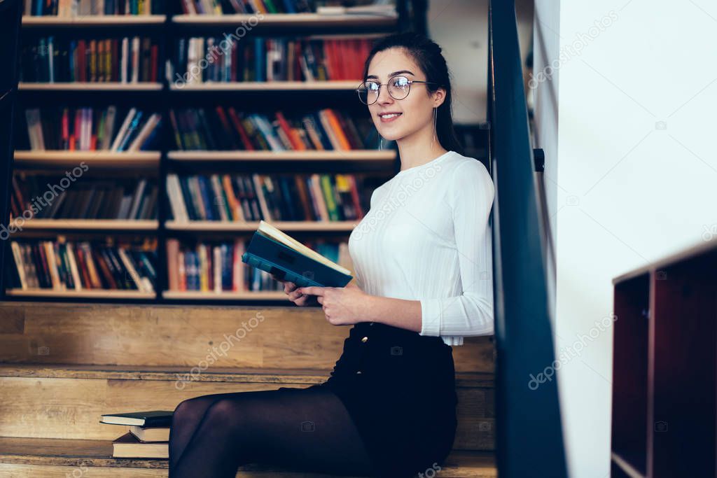 Beautiful young woman in casual wear sitting on stairs with book in college library, attractive intelligent female student looking away dreaming about romantic novel plot spending time on hobby