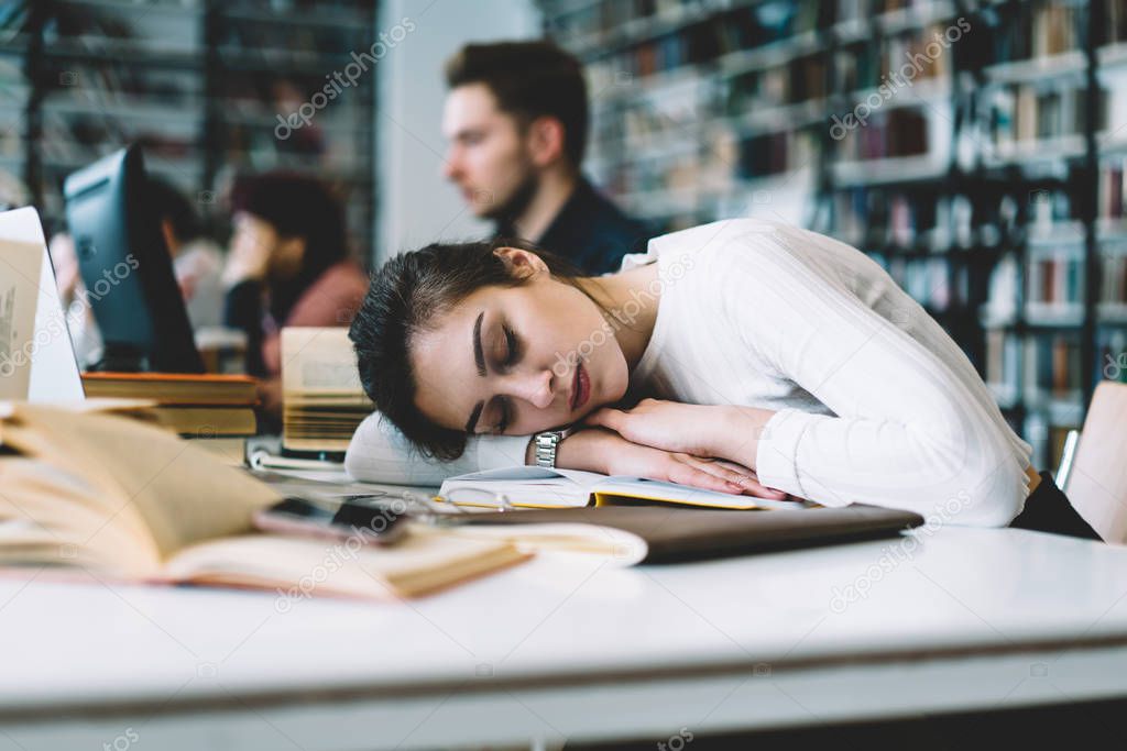 Young female woman sleeping at table with books in university library tired of learning and preparing for examinations, overworked female student need rest concept lying on desktop bored of studyin