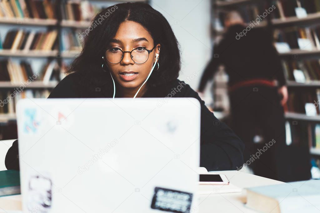 Young african american female freelancer in eyewear for vision protection working on laptop computer, concentrated dark skinned student watching tutorial online learning online course in campu