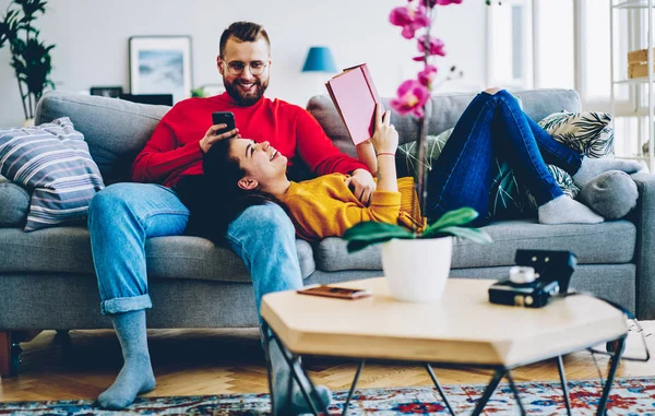 Cheerful couple in love feeling happiness with spending leisure together at home on weekends, man using smartphone and wifi at house while his girlfriend reading book and joking during conversation