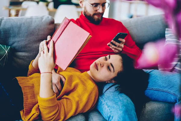 Young family spending time at comfortable apartment together, hipster girl lying on sofa with boyfriend making online booking via smartphone connected to wifi while reading interesting literature