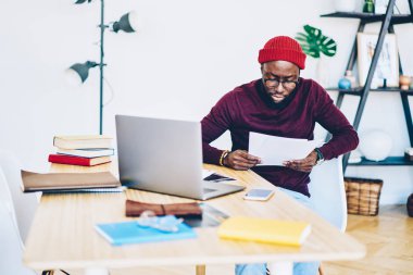 Dalgın koyu tenli erkek freelancer bilgilerini deskto oturup rapor okuma gözlüğü ev iç, ciddi Afro-Amerikan hipster adam uzak iş yaptığını kağıtları üzerinden kontrol