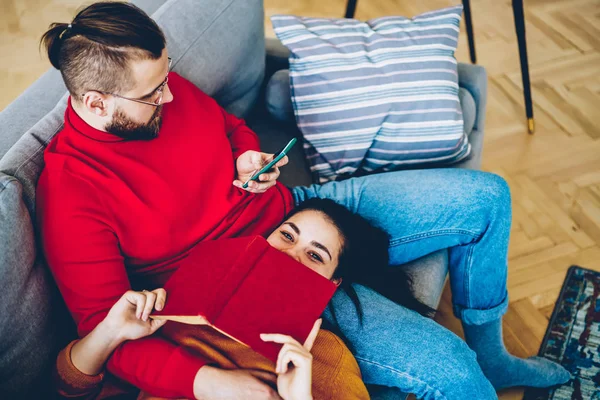 Happy young woman spending time with her husband checking notification on smartphone while sitting on sofa, hipster girl enjoying free time with interesting book at home interior