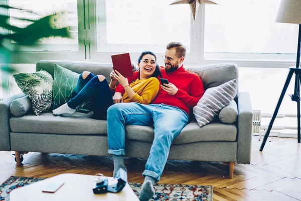 Happy emotional couple laughing at funny content in social networks enjoying free time together , smiling woman lying with book on sofa next to her boyfriend talking and joking