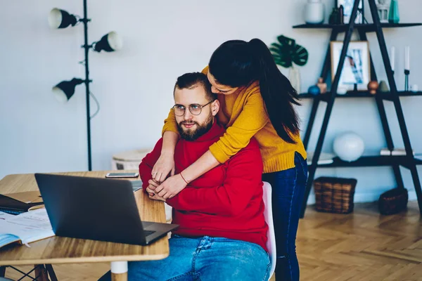 Woman hugging her boyfriend while he working remotely on laptop computer at home interior, irritated male feeling nervous about girlfriend interrupting his distance job process at apartmen