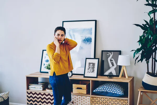 Serious caucasian woman pondering during telephone conversation with service operator booking delivery at home, young hipster girl making mobile phone call standing in modern interior apartmen