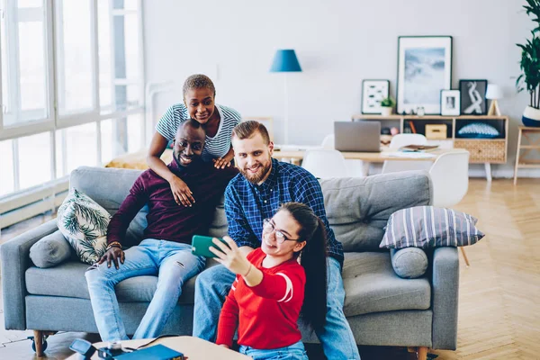 Group of four multiracial male and female friends posing for common selfie on smartphone camera resting at home interior,hipsters sitting on couch at apartment making picture on mobile phone