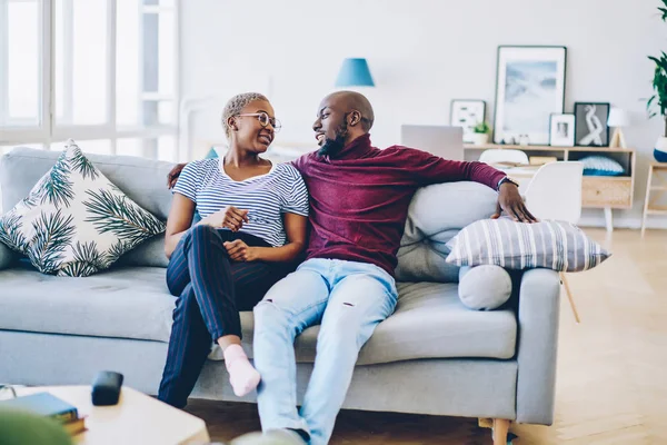 Positive african american young marriage talking to each other sitting on comfortable couch, dark skinned romantic couple enjoying free time together communicating at cozy interior in living room