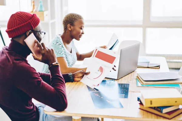 Back view of african american male talking on mobile phone about graphics sitting net to female colleague  searching information on netbook, skilled dark skinned employees making research for design