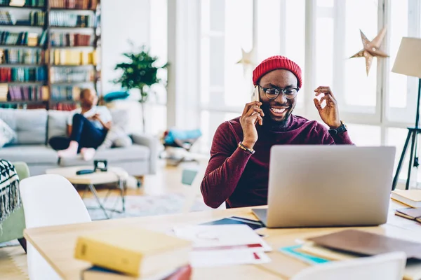 Cheerful dark skinned male freelancer satisfied with getting good news talking on mobile phone, happy african american hipster guy excited with completed online project communicating on cellula