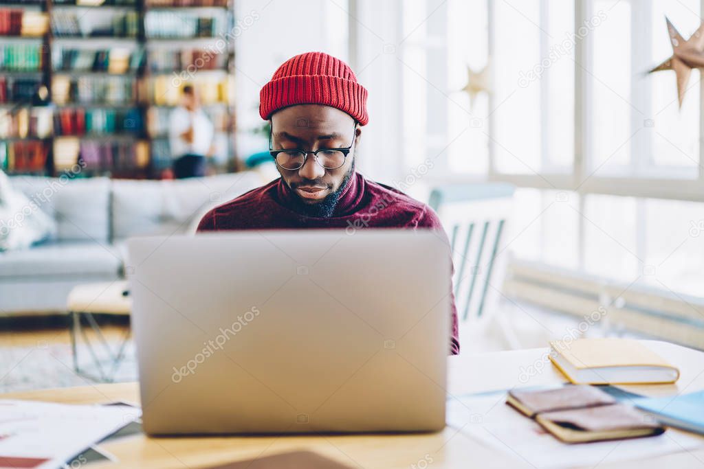Serious african american hipster guy in spectacles typing on laptop computer sitting at home interior, dark skinned male blogger working remotely concentrated on keyboarding publication for blo