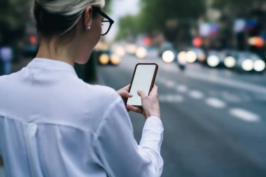 Arkadan görünüm smartphone aygıt tutarak ve app ayakta bulanık arka plan üzerinde kentsel ortamda yönde ara cihazın boş ekranda parmakları ile dokunarak hipster Blogger kırpılmış