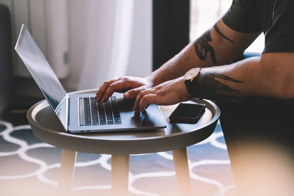 Cropped view of man\'s hands with cool tattoo keyboarding information on modern laptop device using wireless internet connection.Hipster student searching application on web page on digital netbook