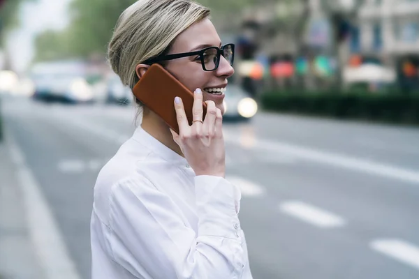 Side view of positive hipster female blogger laughing during phone conversation on smartphone device in urban setting.Cheerful businesswoman calling on telephone and talking with friend in roaming